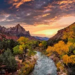 De kartelige rotsformatie The Watchman kleurt oranje bij zonsondergang boven een kronkelende rivier omzoomd door herfstbomen in Zion National Park