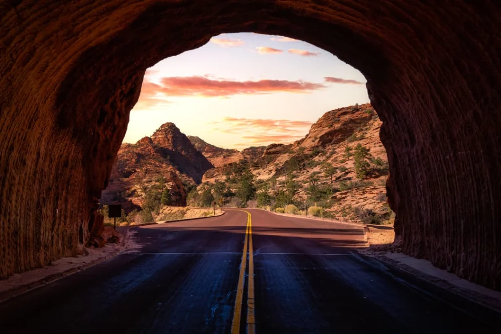 Weg door rode rotsformaties gezien vanuit de opening van een tunnel, met een gekleurde avondlucht in Zion National Park, Utah