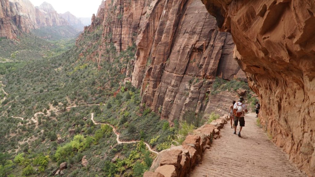 Wandelaars lopen over een smal pad tegen een rode rotswand met uitzicht op de groene vallei van Zion National Park