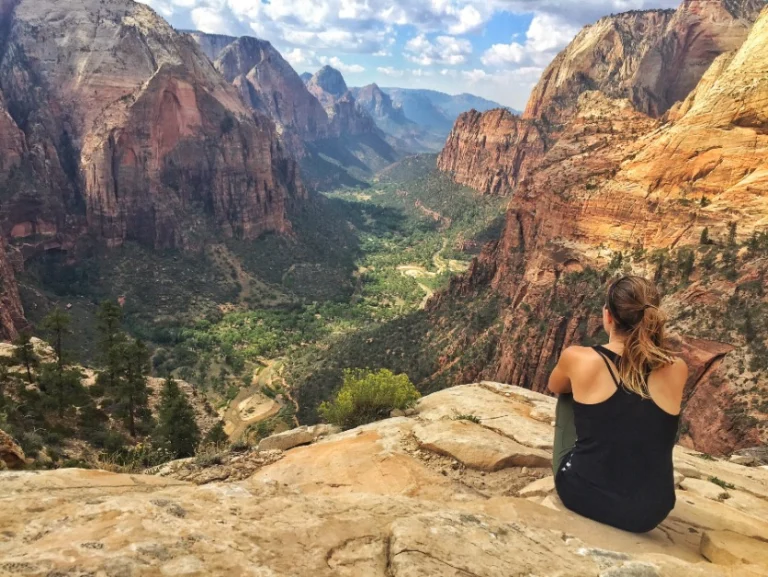 Een vrouw zit op een rotsplateau en kijkt uit over een groen dal tussen rode rotswanden in Zion National Park