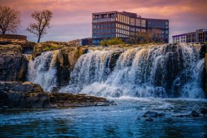 Brede waterval stroomt over rotsblokken in Falls Park in Sioux Falls met een verlicht gebouw op de achtergrond bij zonsondergang