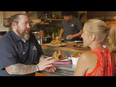 Een getatoeëerde chef met baard laat een bord eten zien aan een lachende vrouw terwijl op de achtergrond een kok in de keuken werkt