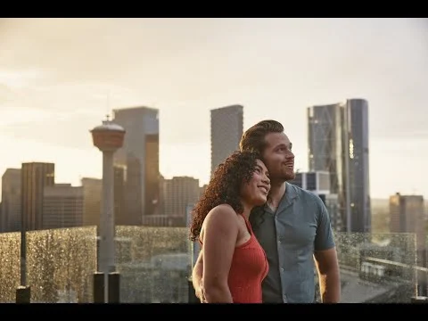 Een lachend koppel staat op een dakterras met uitzicht op de skyline van Calgary tijdens zonsondergang, met de Calgary Tower op de achtergrond