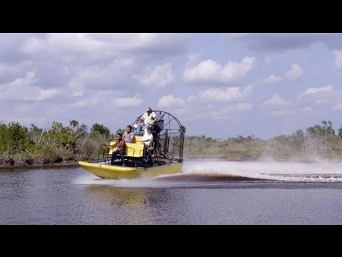 Een gele airboat met passagiers en gids raast door het moerasgebied van Everglades National Park onder een bewolkte lucht