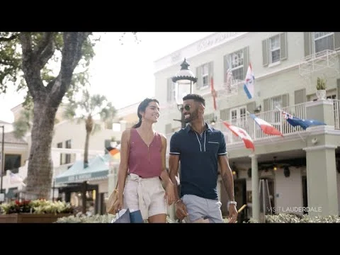 Een lachend koppel loopt gearmd door een winkelstraat in Fort Lauderdale met palmbomen en vlaggen op de achtergrond