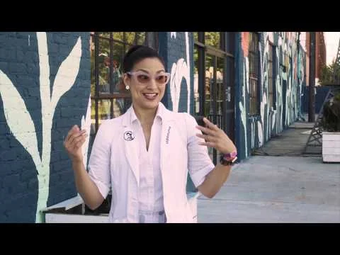 Een vrouw met zonnebril en witte blazer praat en gebaart voor een beschilderde bakstenen muur in een straat in Georgia