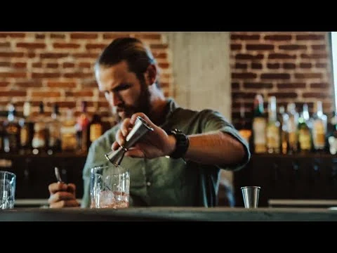 Een bebaarde bartender in een bar in Honolulu schenkt met een maatbeker drank in een glas met ijs