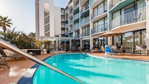 Rond zwembad met turquoise water omringd door ligstoelen en parasols voor een hotelgebouw met balkons