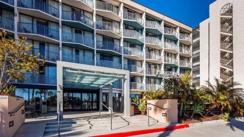 Meerdere verdiepingen met balkons van een hotel, met een overdekte ingang en palmbomen op de voorgrond