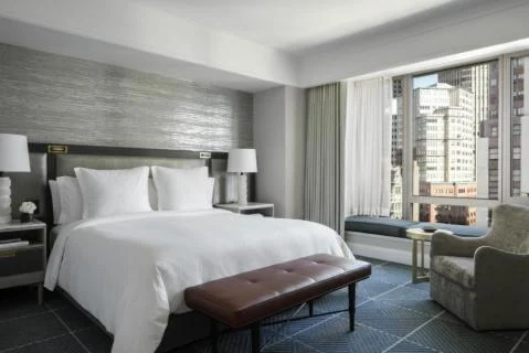 Hotelkamer met een groot wit bed, een zitbank aan het voeteneind en een raam met uitzicht op wolkenkrabbers