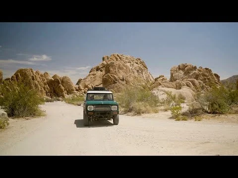 Groene terreinwagen rijdt over een zandpad tussen grote rotsformaties onder een strakblauwe lucht in Joshua Tree National Park