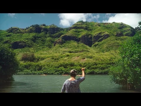 Een vrouw staat bij een rivier op Kauai met op de achtergrond weelderige groene bergen en kajakkers op het water