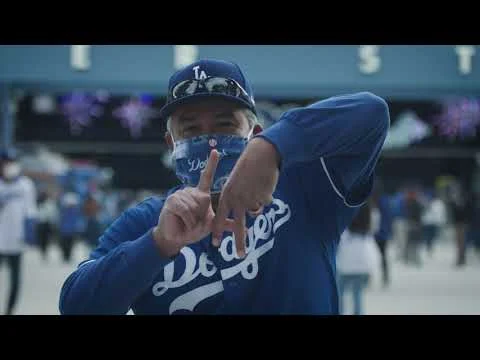 Man met een Dodgers pet, shirt en mondkapje maakt een victoryteken met zijn vingers bij het stadion