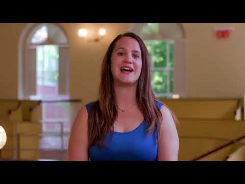 Videobeeld van een lachende vrouw die praat in een lichte historische zaal met houten kerkbanken en gewelfde ramen in Massachusetts