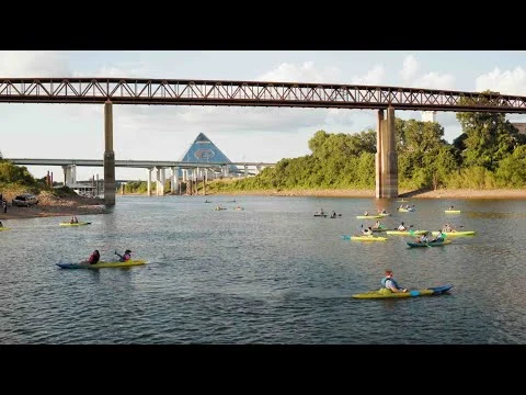 Groep kajakkers vaart over een brede rivier in Memphis, met een spoorbrug en de piramide op de achtergrond