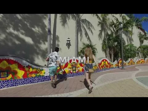 Twee wandelaars lopen over een zonnig pad langs een kleurrijke mozaiekmuur met palmbomen in Miami