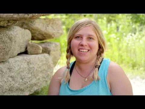Videobeeld van een lachende vrouw met vlechten die praat voor een stenen muur tussen het groen in New Hampshire