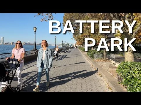 Videostill van twee vrouwen die wandelen op een pad in Battery Park New York langs het water, met de tekst Battery Park in beeld