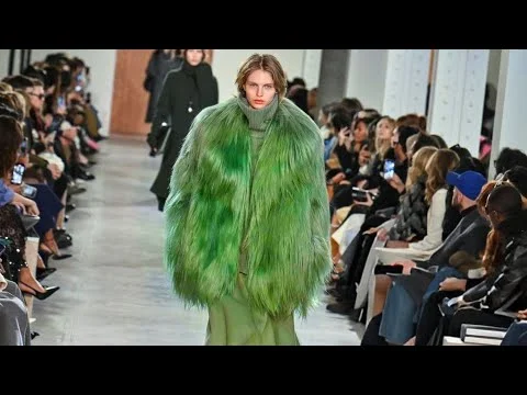 Videothumbnail van een model in een felgroene bontjas dat over de catwalk loopt langs zittend publiek