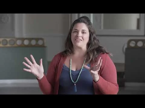 Videostilstaand beeld van een vrouw met een rood vest en een blauwe ketting die gebarend praat in een interieur met gedecoreerde randen