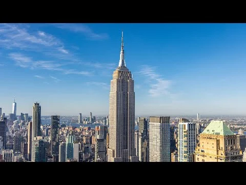 Videothumbnail die de skyline van Manhattan toont met het Empire State Building centraal tegen een heldere blauwe lucht