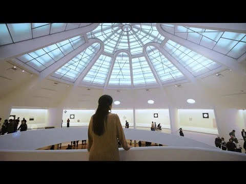 Videothumbnail van een bezoeker die de spiraalvormige binnenkant van het Guggenheim Museum bekijkt onder de koepel met glazen dak