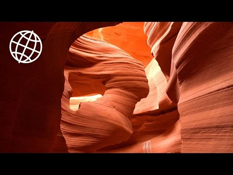 Videostilstaand beeld van een lichtgevende opening tussen de golvende oranje wanden van Antelope Canyon met een wereldbol icoon in de hoek
