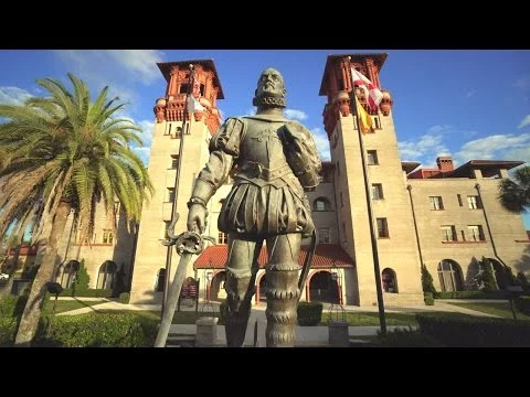 Bronzen standbeeld van een conquistador met zwaard voor een torengebouw met Spaanse vlaggen in Saint Augustine, met palmbomen en blauwe lucht op de achtergrond