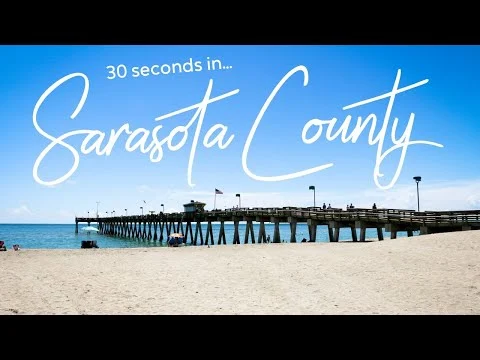 Lange houten pier met vlaggenmasten strekt zich uit over het witte strand van Sarasota County onder een strakblauwe lucht