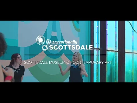 Stilstaand videobeeld van twee vrouwen die praten voor een kleurrijke muur, met tekst over het museum voor hedendaagse kunst
