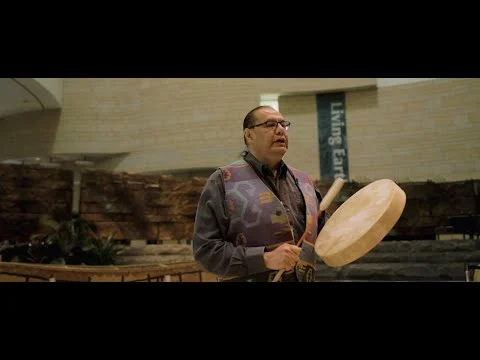 Videothumbnail met een inheemse man die op een handtrommel speelt in een zaal met de tekst Living Earth