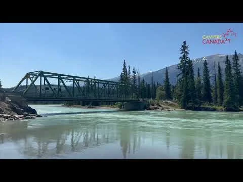 Videothumbnail met een stalen brug over een rivier, dennenbos en besneeuwde bergen op de achtergrond, met het logo Campers Canada