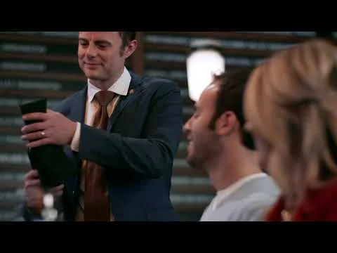 Videostill van een lachende man in kostuum met een glas in zijn hand naast bezoekers aan een tafel in een pub in Washington
