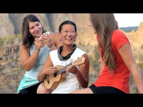 Videostill van drie lachende vrouwen waarvan er een ukelele speelt, met op de achtergrond rotsformaties die lijken op Smith Rock in Oregon