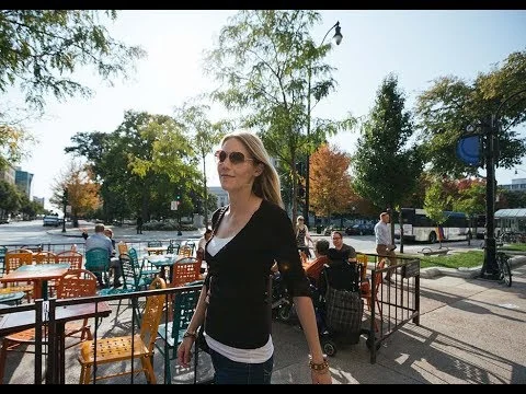 Videobeeld van een vrouw met zonnebril die langs een buitenterras met stoelen en tafels loopt in een stad in Wisconsin