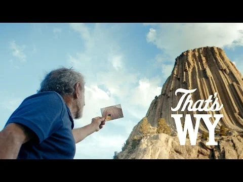 Grijze man houdt een oude foto omhoog tegenover de rotswand van Devils Tower met de tekst That's WY in beeld