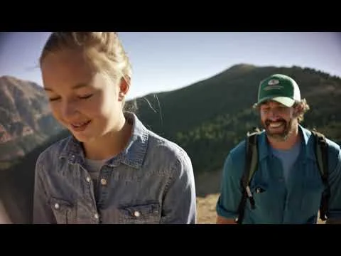 Lachend meisje op de voorgrond met een man met rugzak en pet achter haar tijdens een wandeling in bergachtig landschap