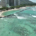Luchtfoto van hotels en palmbomen langs het witte strand van Waikiki Beach met zwemmers in turquoise water en de Diamond Head op de achtergrond