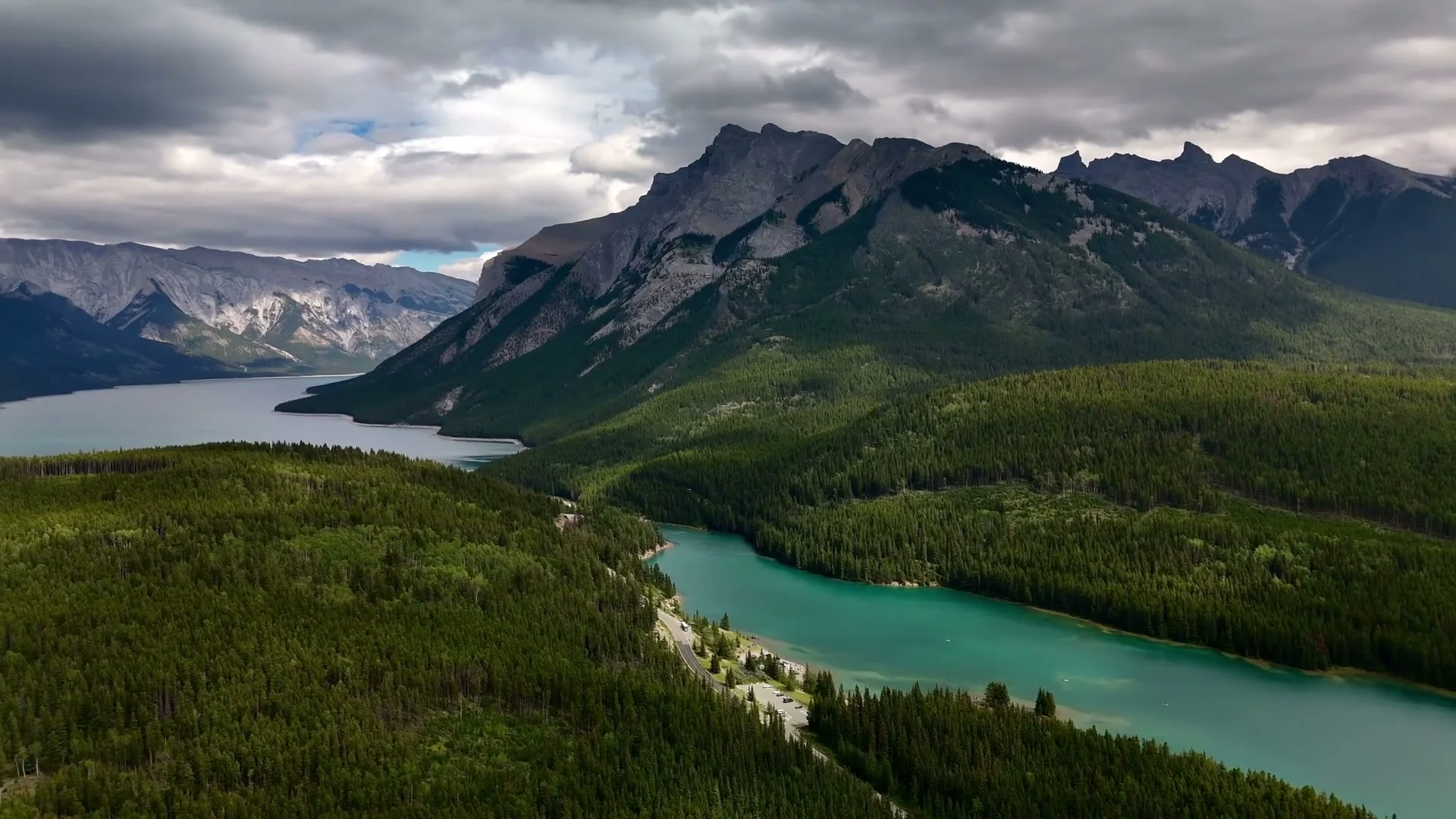 Luchtfoto van een turquoise meer omringd door dennenbossen en bergtoppen in de Canadese Rockies onder een bewolkte lucht