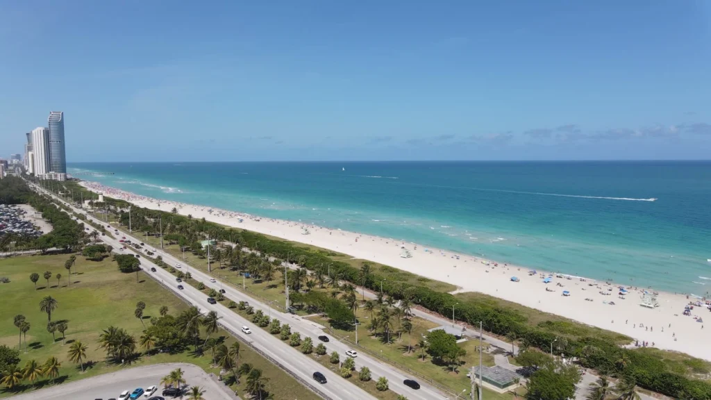 Luchtfoto van een lang wit strand met turquoise zee, een boulevard met palmbomen en een hoge woontoren in Miami Beach