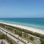 Luchtfoto van een lang wit strand met turquoise zee, een boulevard met palmbomen en een hoge woontoren in Miami Beach