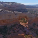 Grote natuurlijke boog van rode zandsteen torent boven een uitgestrekte canyon met struiken en verre bergen